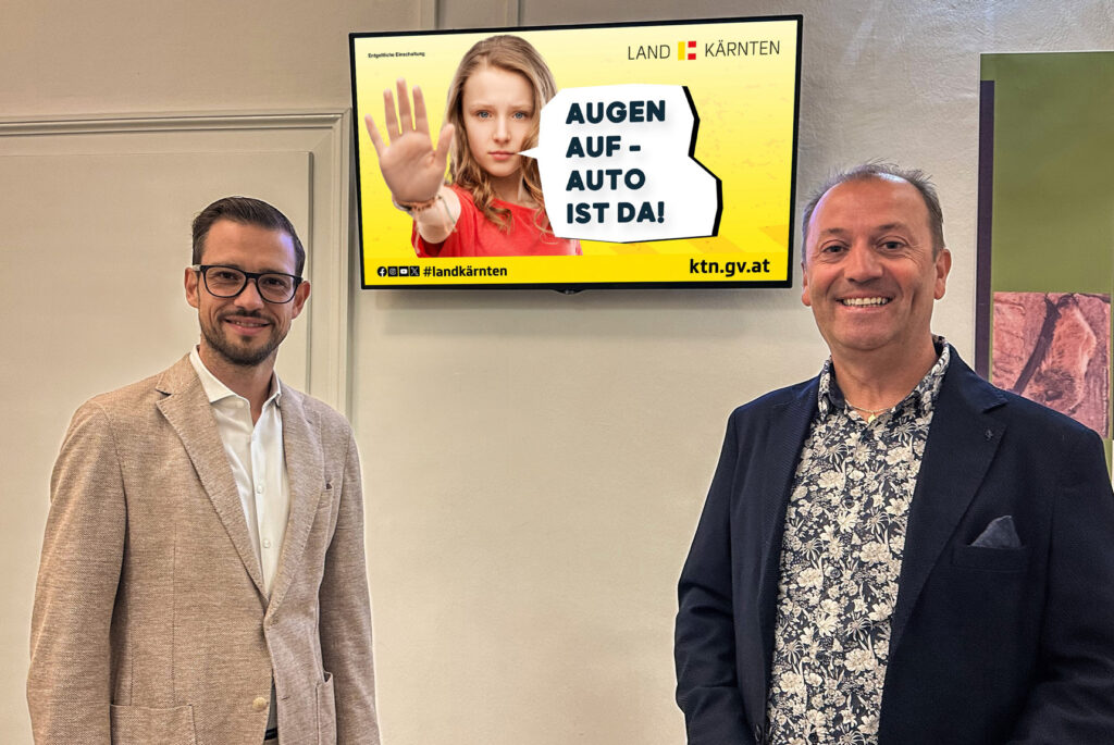 LR Sebas­ti­an Schu­sch­nig und Andre­as Lan­ner vor einem Indoor Screen. Zu sehen am Screen eine Grafik mit dem Titel "Augen auf, Auto ist da!"